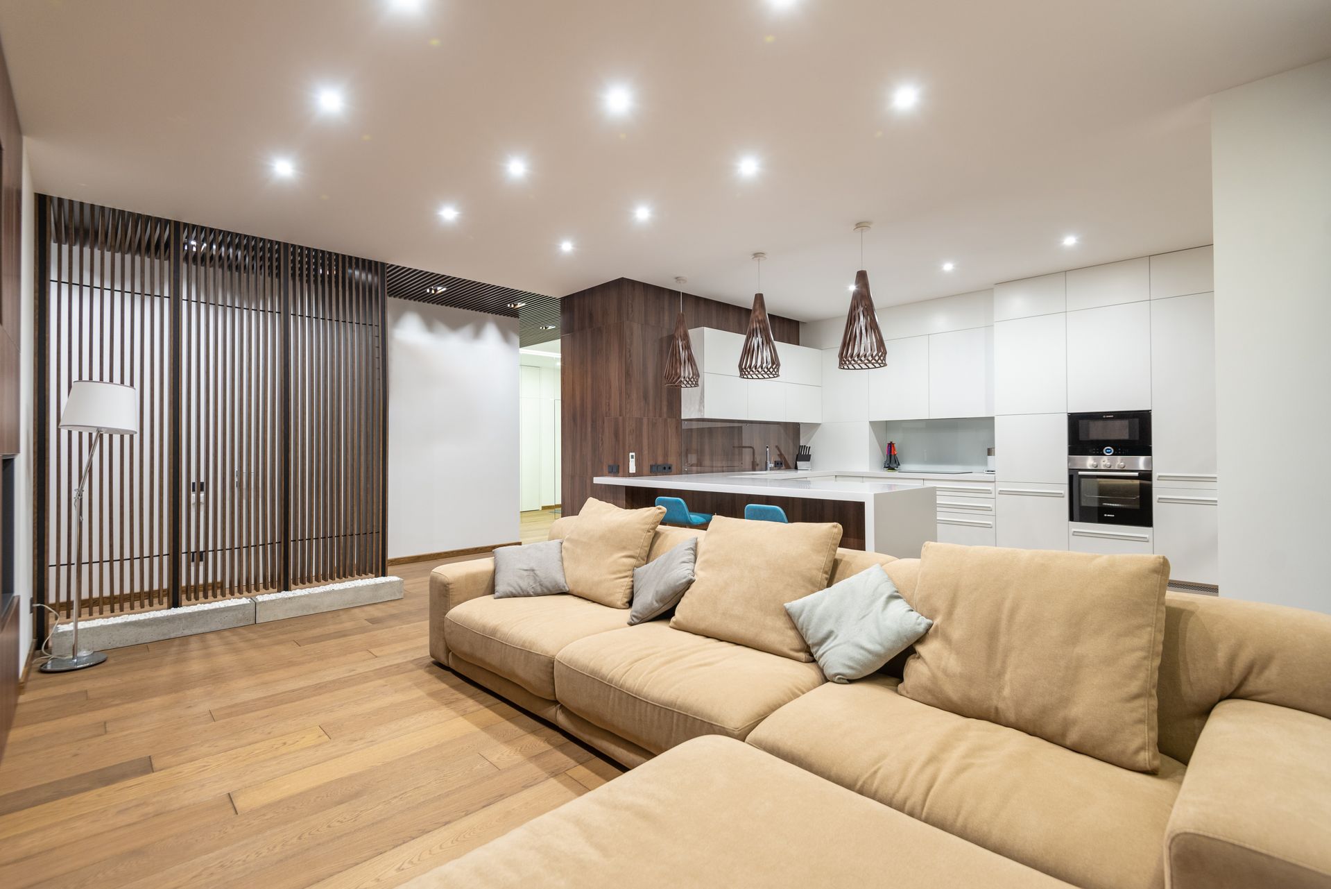 A beige sectional sofa in a living room facing an open kitchen with white cabinets, a central island, and wood-paneled walls.