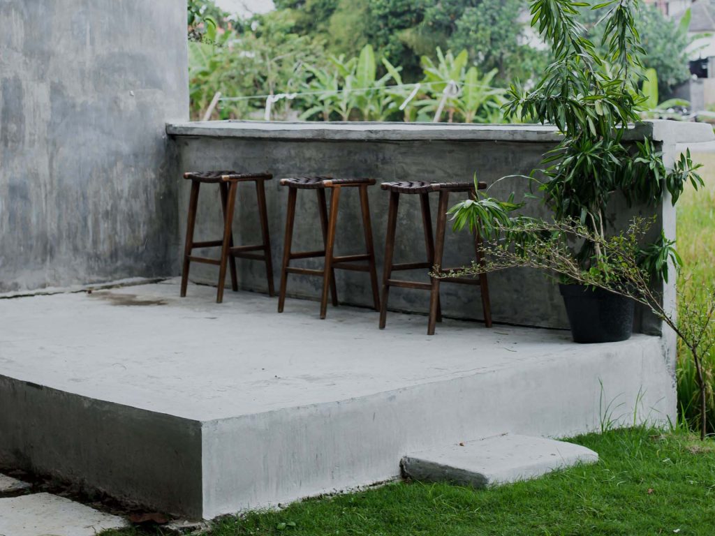 Three wooden bar stools lined up against a concrete outdoor counter with a potted plant nearby.