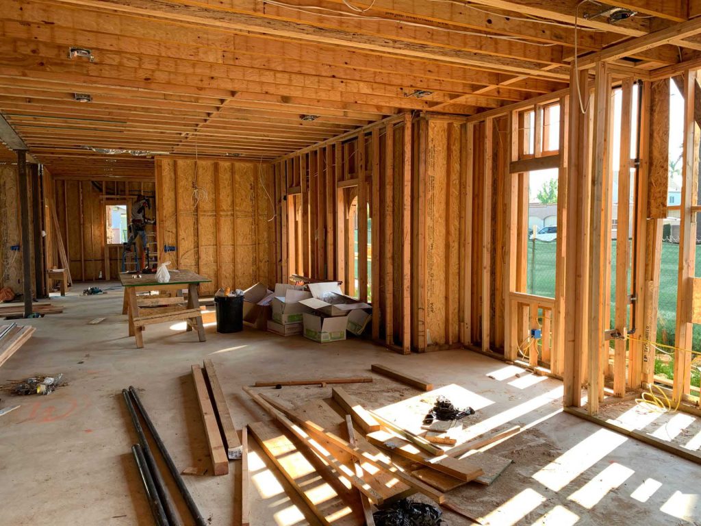 Interior of a house under construction with exposed wooden wall framing, floor joists, and scattered lumber.