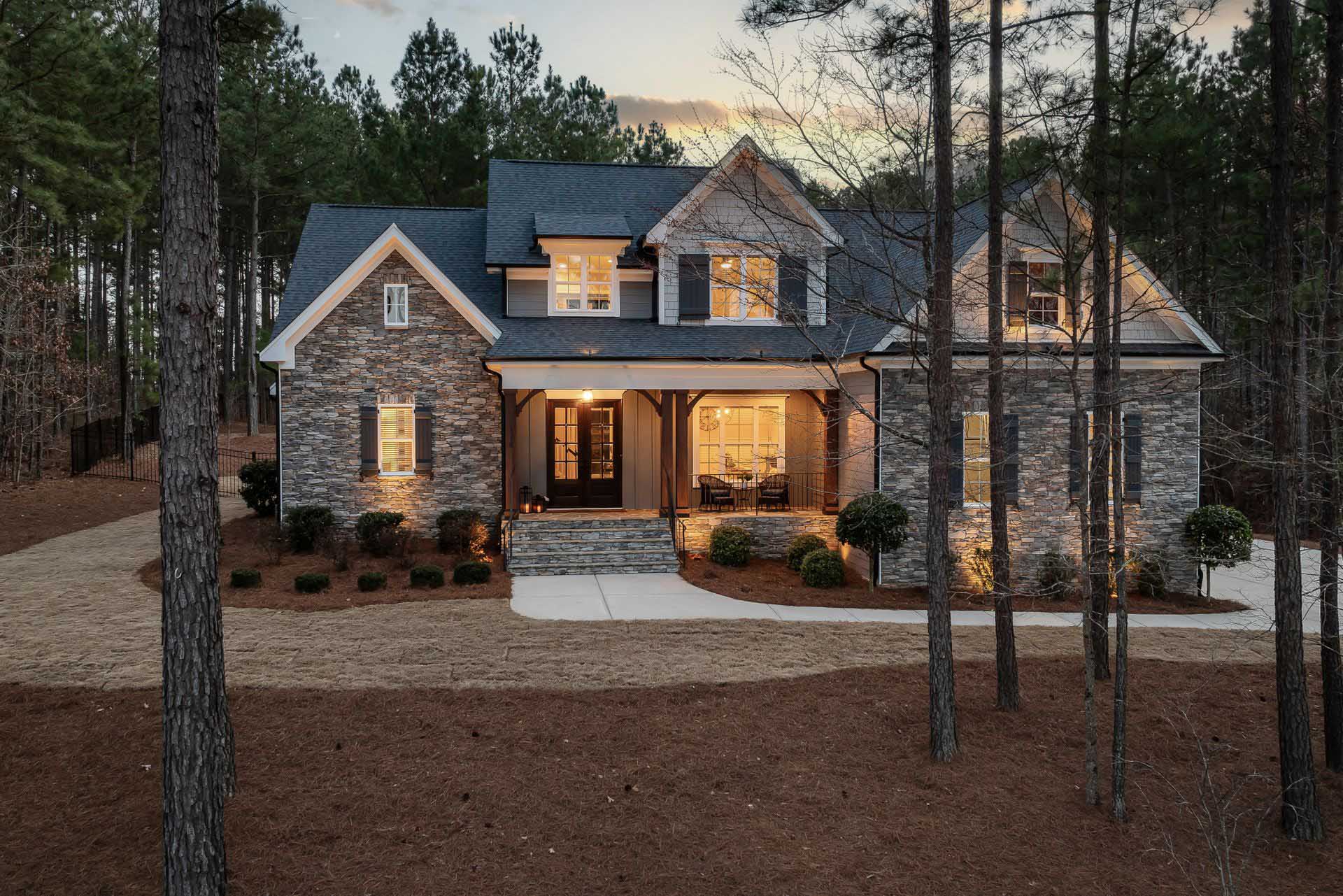 A two-story stone and siding house at dusk, with interior lights glowing and a covered front porch, viewed through a wooded yard.