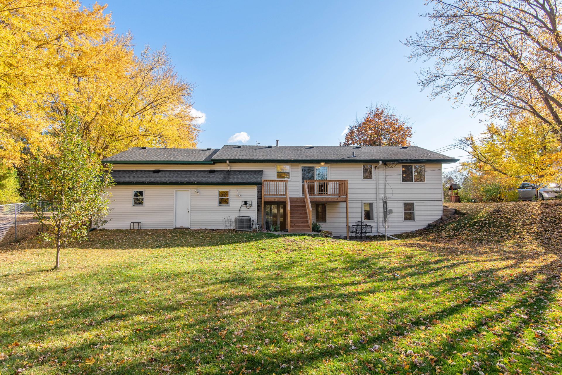 The back exterior of a single-story house with a wooden deck and staircase, surrounded by autumn trees and a grassy yard.