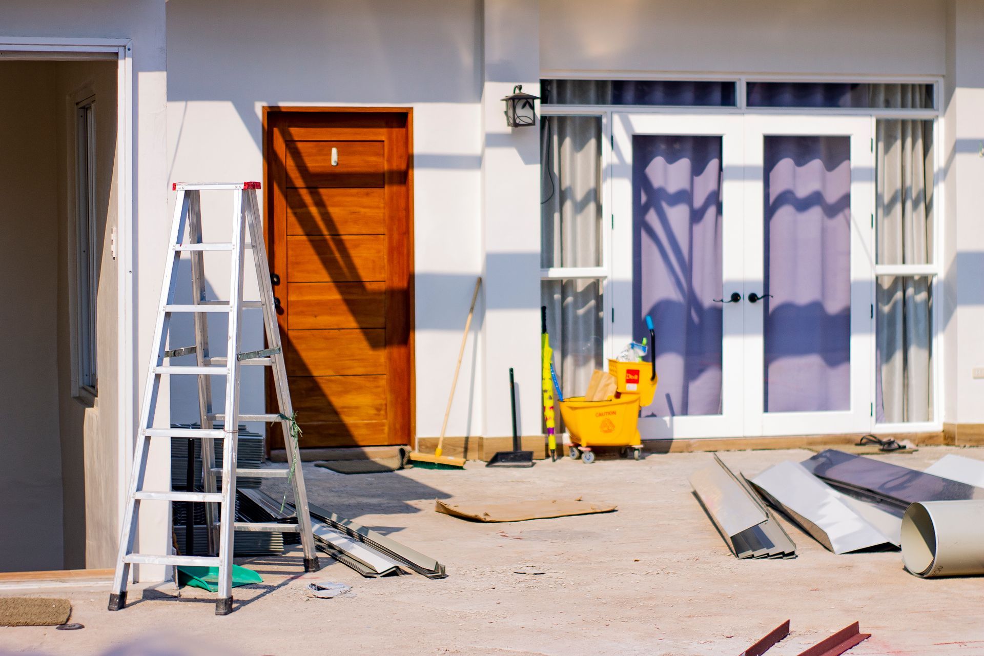 A stepladder, a yellow janitorial cart, and several metal construction panels on a patio in front of a house with a wooden door and glass doors.