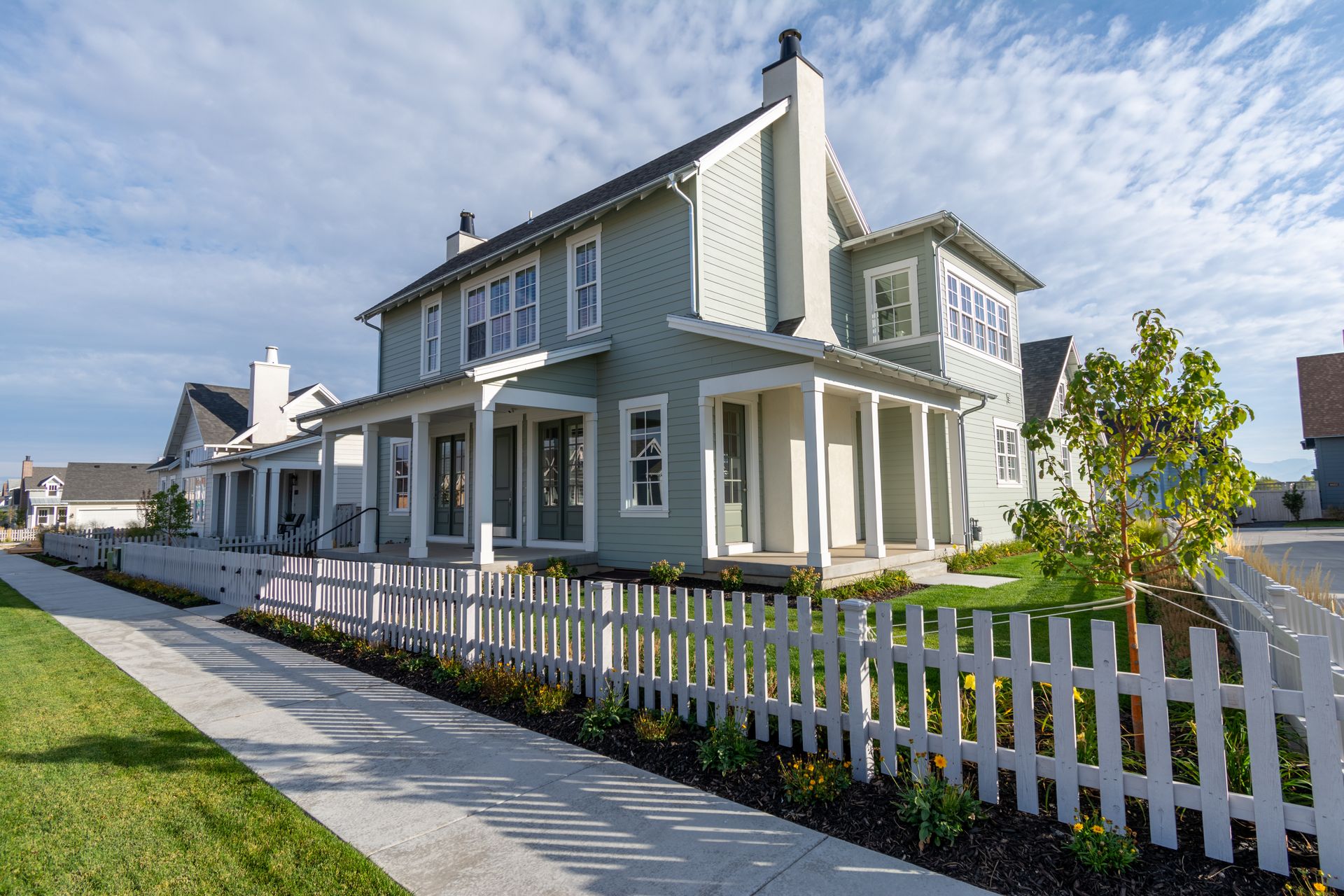 A two-story, light green house with a white picket fence and a covered front porch, seen from a sidewalk.