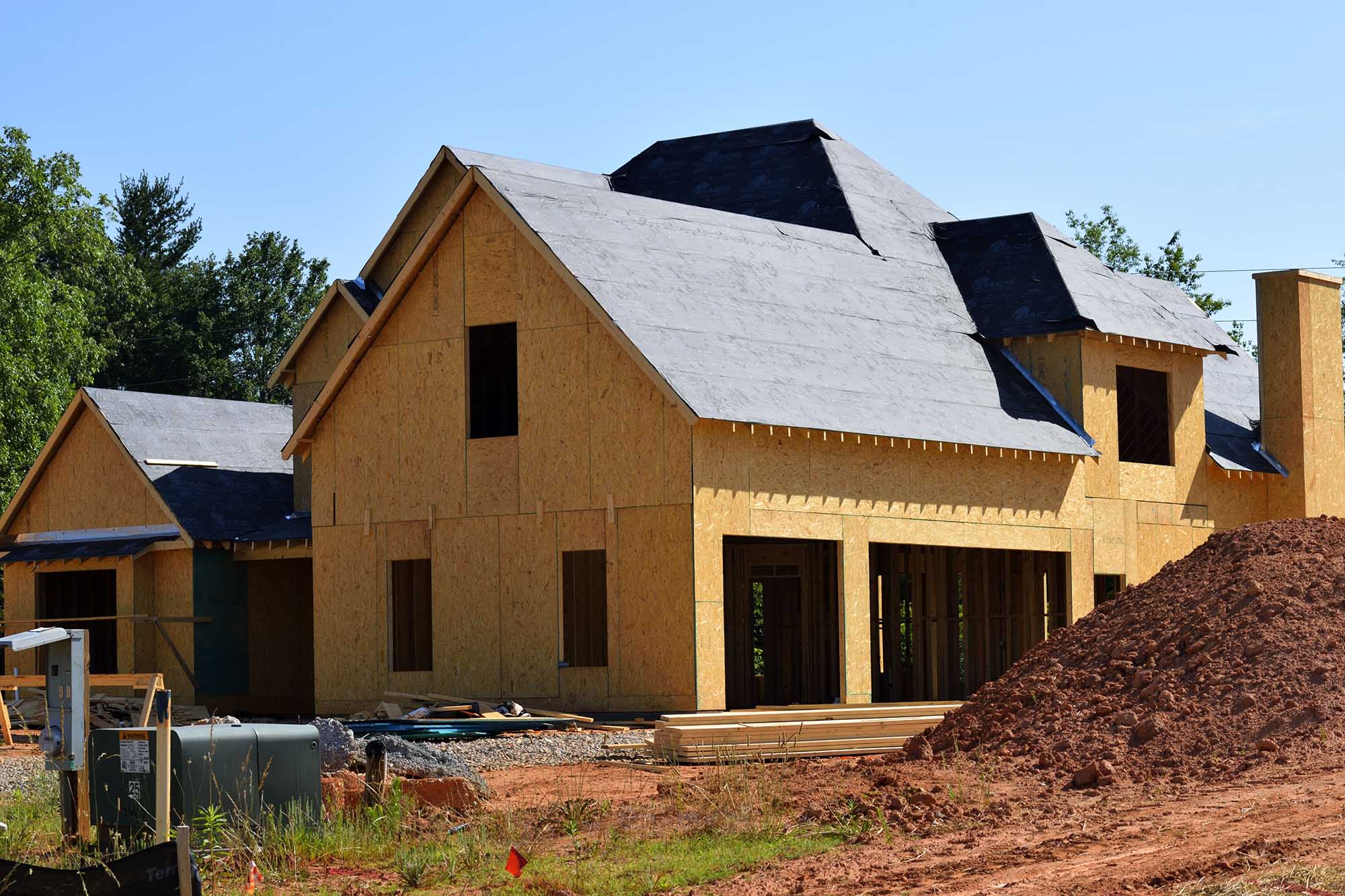 A residential house under construction, featuring exposed plywood walls, a roof covered in black underlayment, and a large pile of dirt in the foreground.
