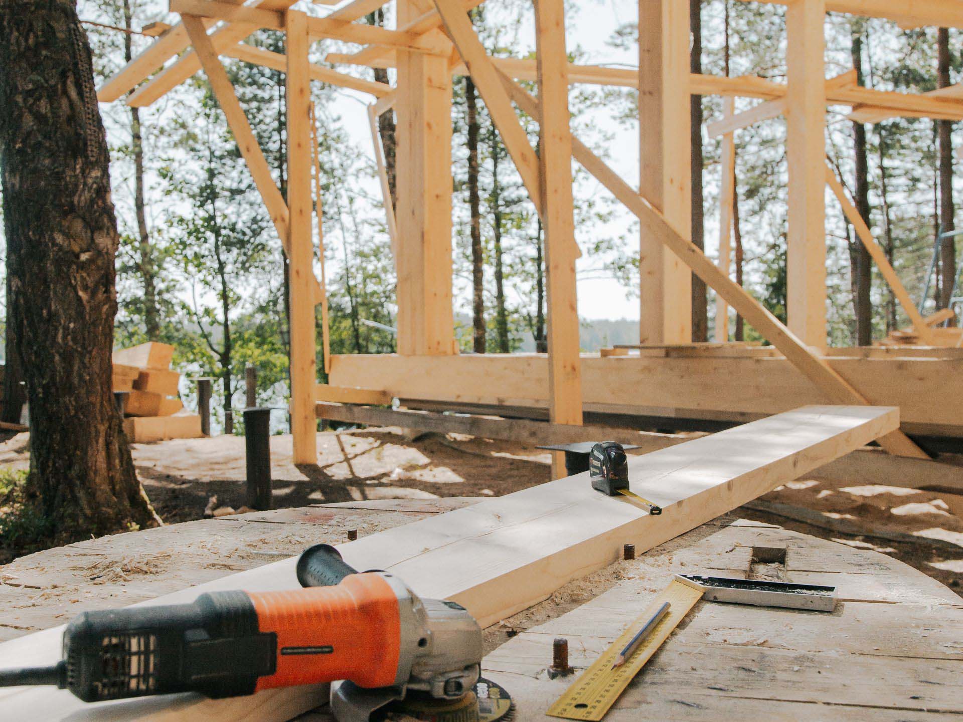 An angle grinder, tape measure, and spirit level sit on a wooden table in front of the timber frame of a building under construction.